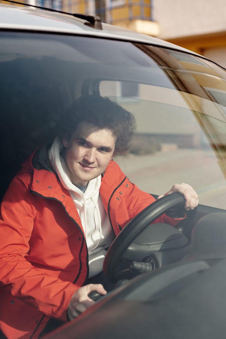 A young adult man smiling while sitting behind the steering wheel of a vehicle. Un jeune homme adulte souriant assis derrière le volant d'un véhicule.