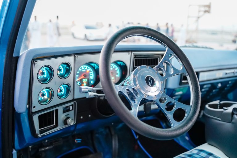 Close-up view of a classic car's dashboard and steering wheel, featuring analog speedometers. Vue rapprochée du tableau de bord et du volant d'une voiture classique, présentant des compteurs de vitesse analogiques.